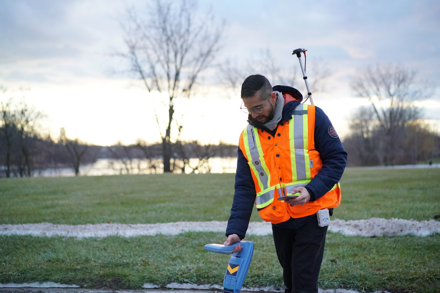 A field technician in a high-visibility vest using a utility locator, with a GNSS antenna on a backpack pole and a phone in hand.